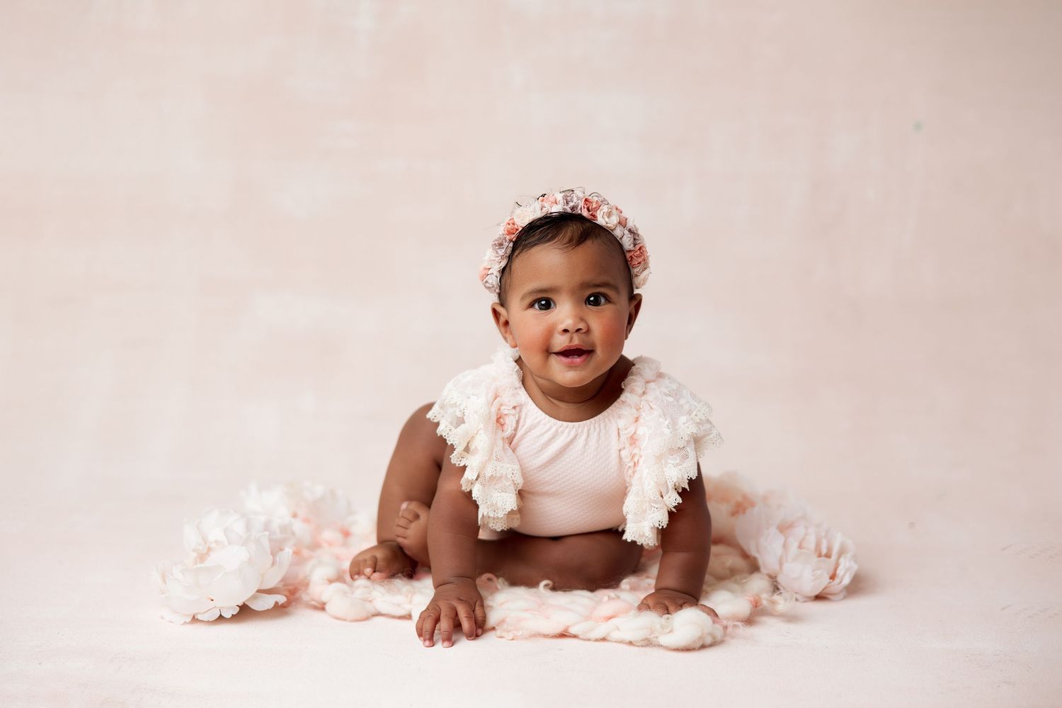 A baby wearing a pink tulle dress and headband poses on a soft pink backdrop during a professional photoshoot.