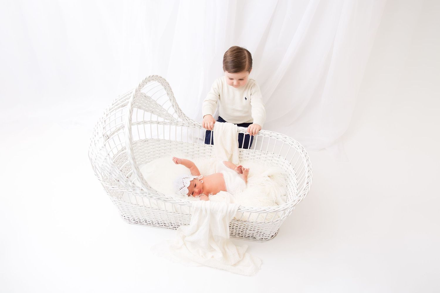 A baby sits in a white vintage wicker bassinet wearing white clothing against a bright white background.