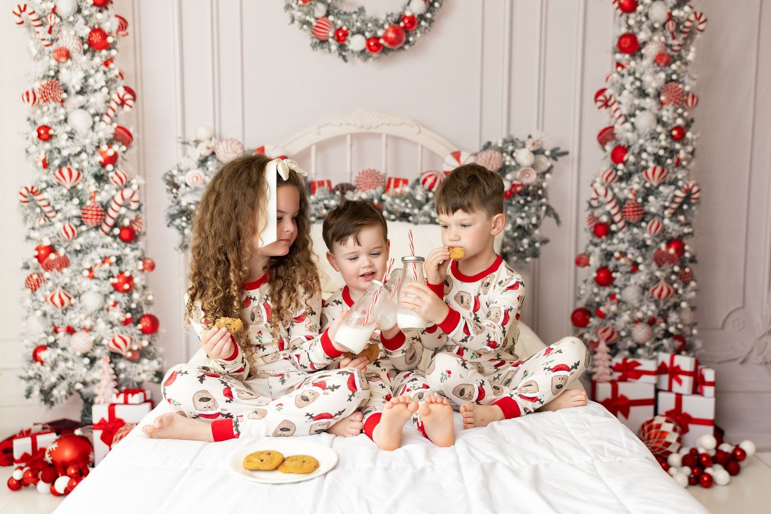 Three children in matching Christmas pajamas sharing cookies on decorated white bed.