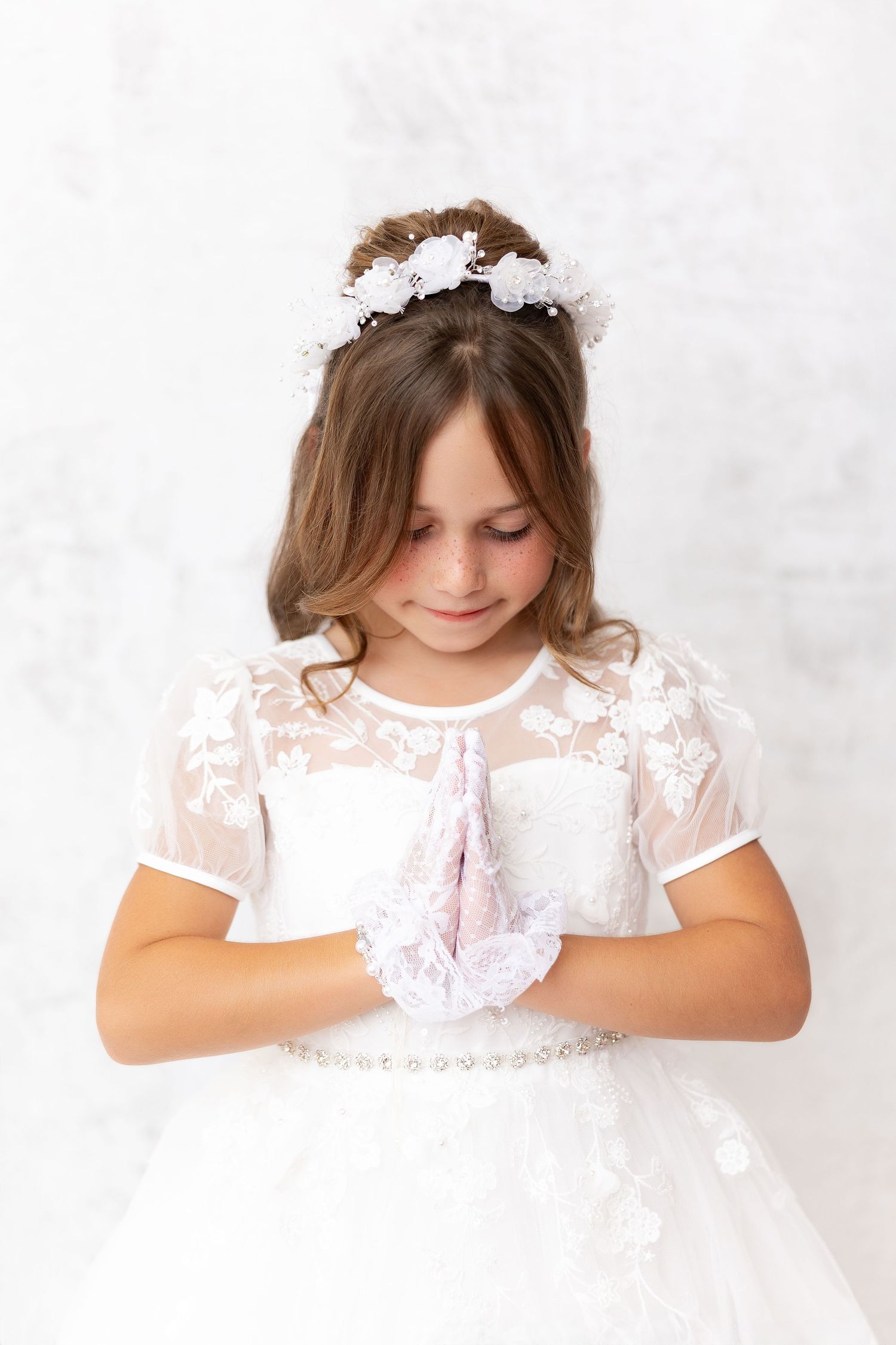 A young child in a white lace first communion dress and gloves looking down with white floral headpiece.