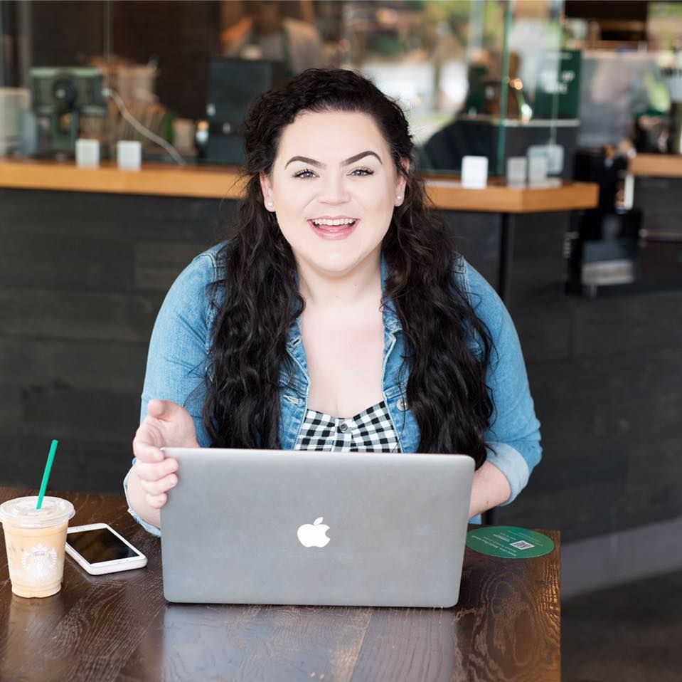 A person in gingham attire and blue cardigan works on MacBook laptop at cafe table with iced coffee nearby.