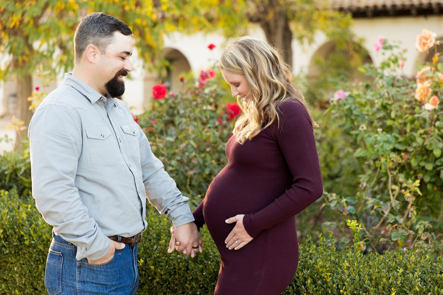 Couple shares intimate moment during maternity photoshoot in garden with roses.