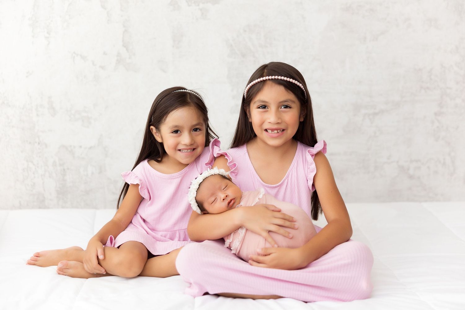 Three siblings in pink dresses sitting together tenderly on white backdrop.