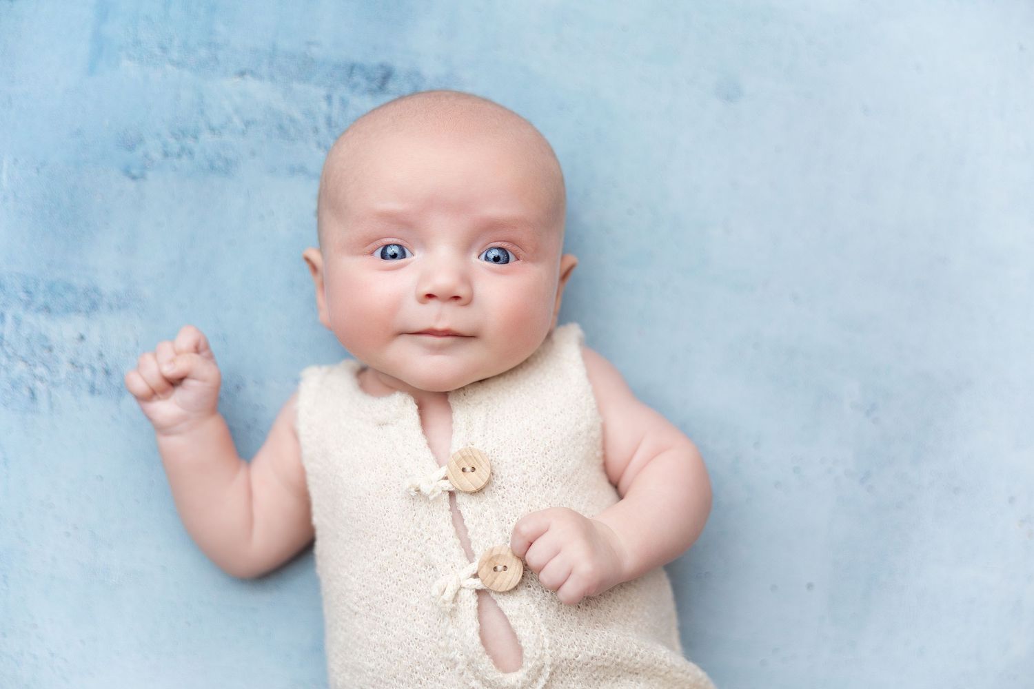 Baby wearing cream-colored sleeveless outfit lying on blue background.