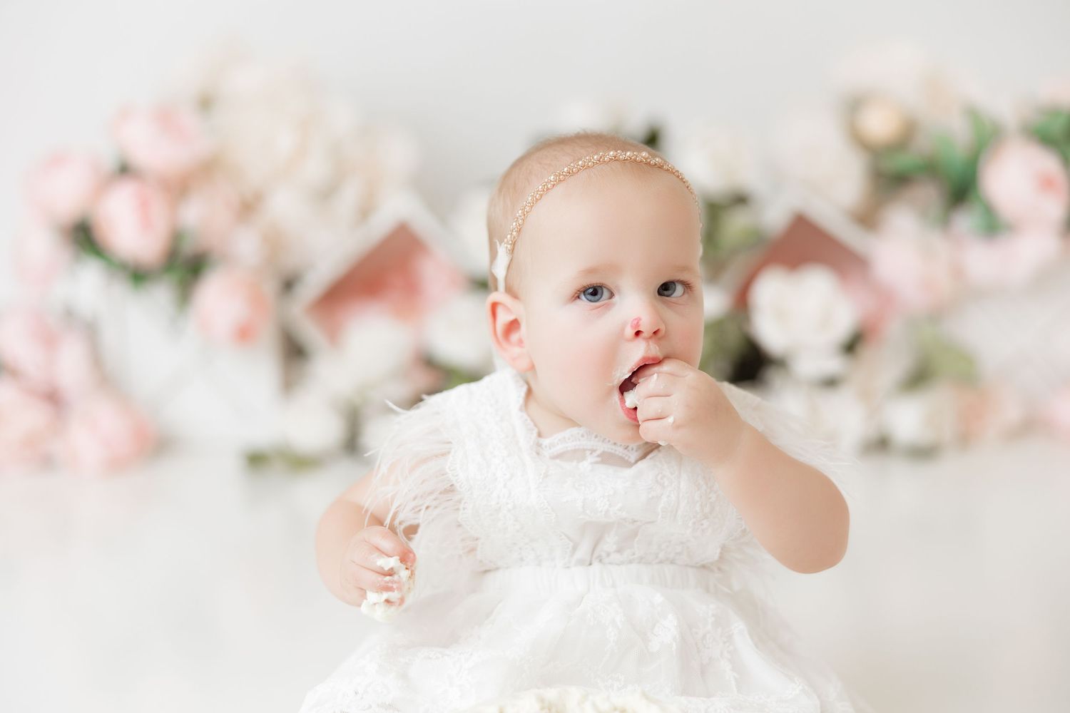Baby in white dress reaching joyfully during cake smash photoshoot with pink flowers.