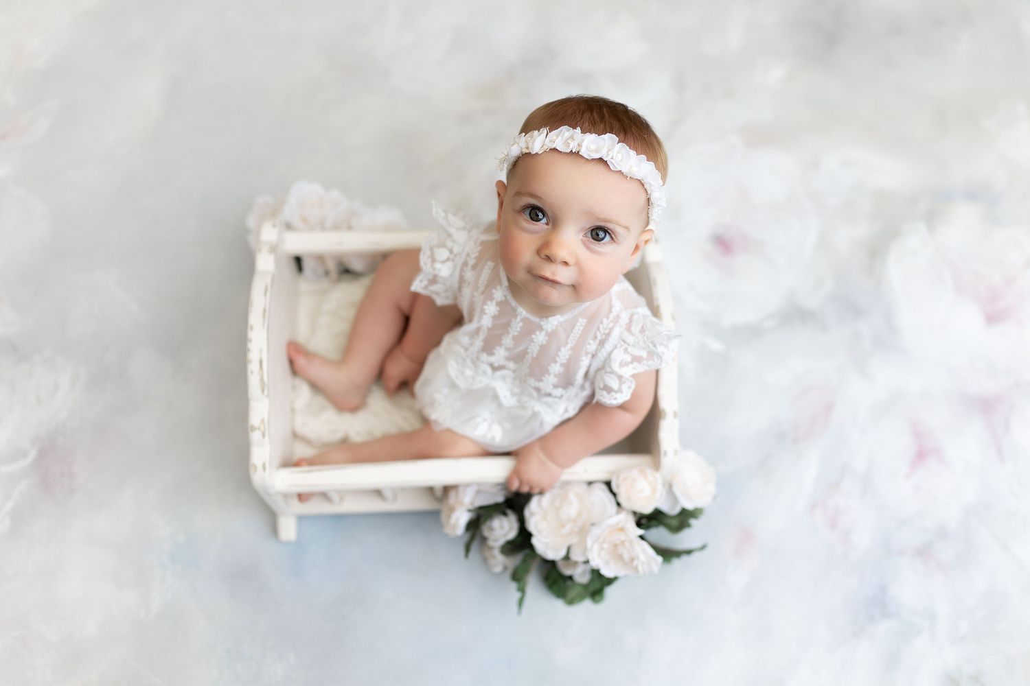Baby in white lace outfit poses on small vintage bed with floral decor.