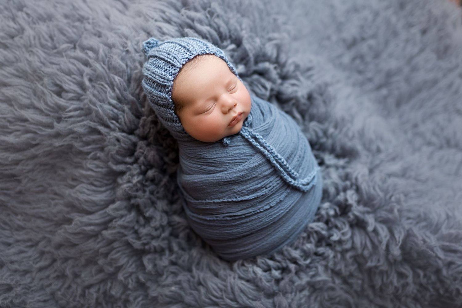 Sleeping newborn wrapped in gray textile on plush gray fur backdrop.