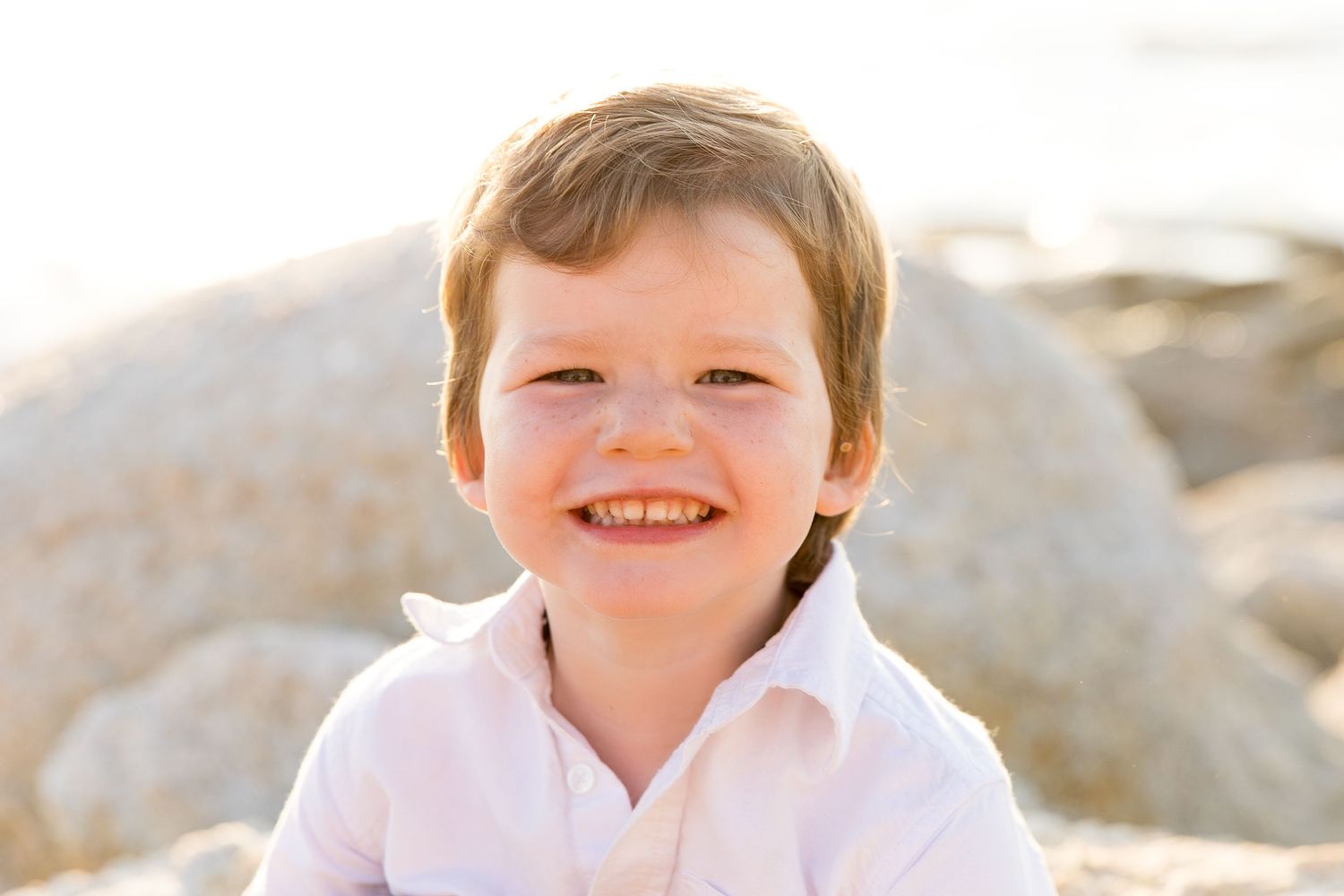 A child in a white polo shirt smiles brightly against a rocky beach backdrop.
