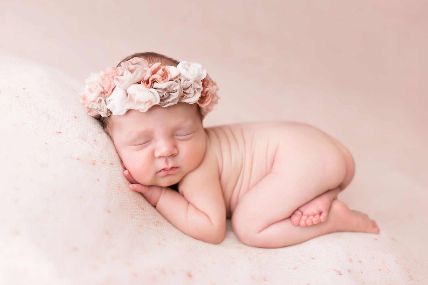 A peaceful sleeping newborn wearing a pink floral crown posed on soft pink fabric.