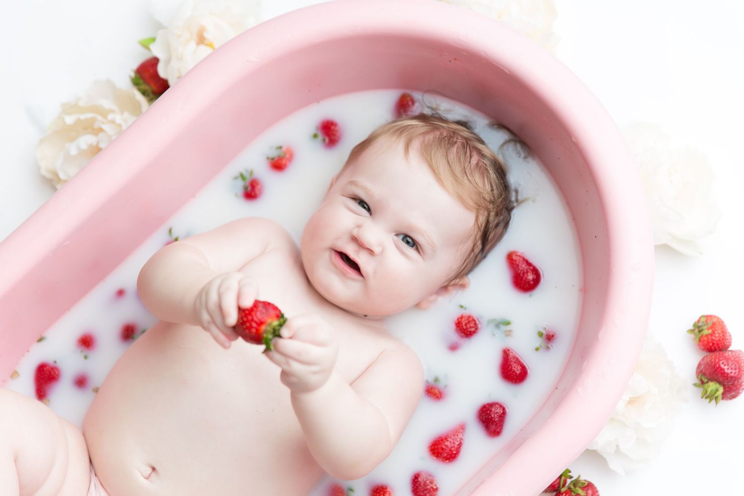 Artistic milk bath photography with baby surrounded by fresh strawberries and flowers.