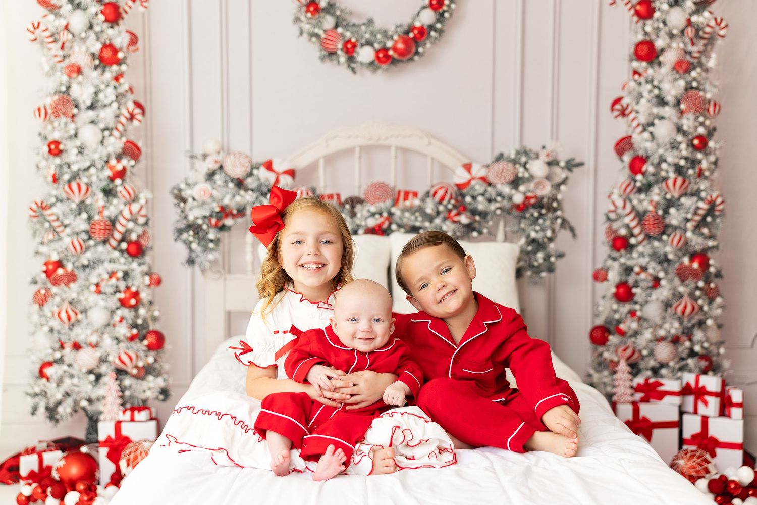 Three people in red holiday pajamas sitting together on white bed with Christmas decorations.