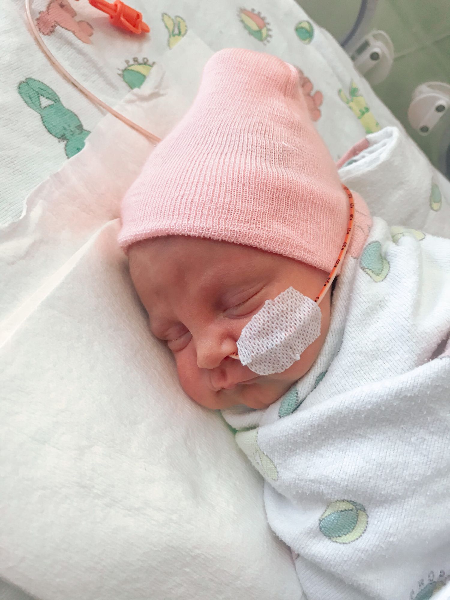 A sleeping newborn wearing a pink knit hat and medical eye patch rests on patterned bedding.