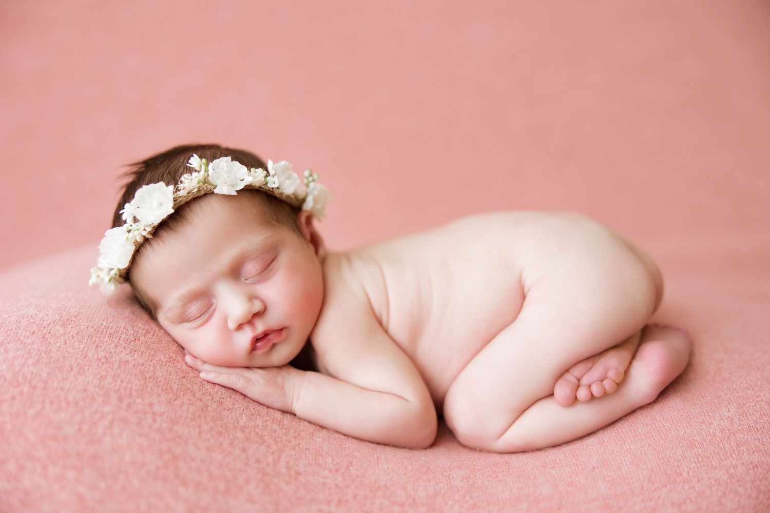 Sleeping newborn baby wearing white flower crown lying on soft pink backdrop in artistic studio portrait.