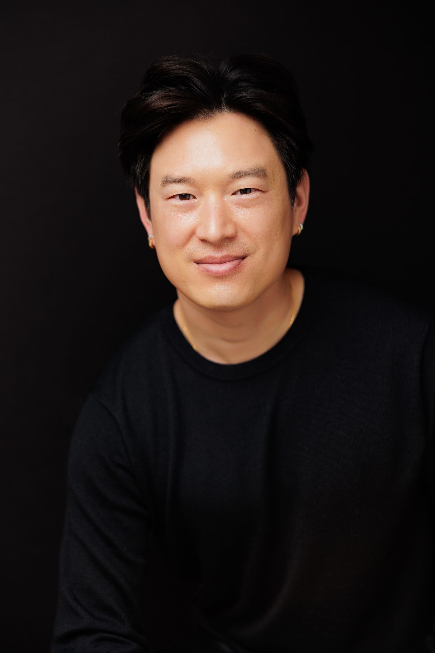 Smiling Asian man in black long-sleeve shirt poses against dark background in professional headshot photo.