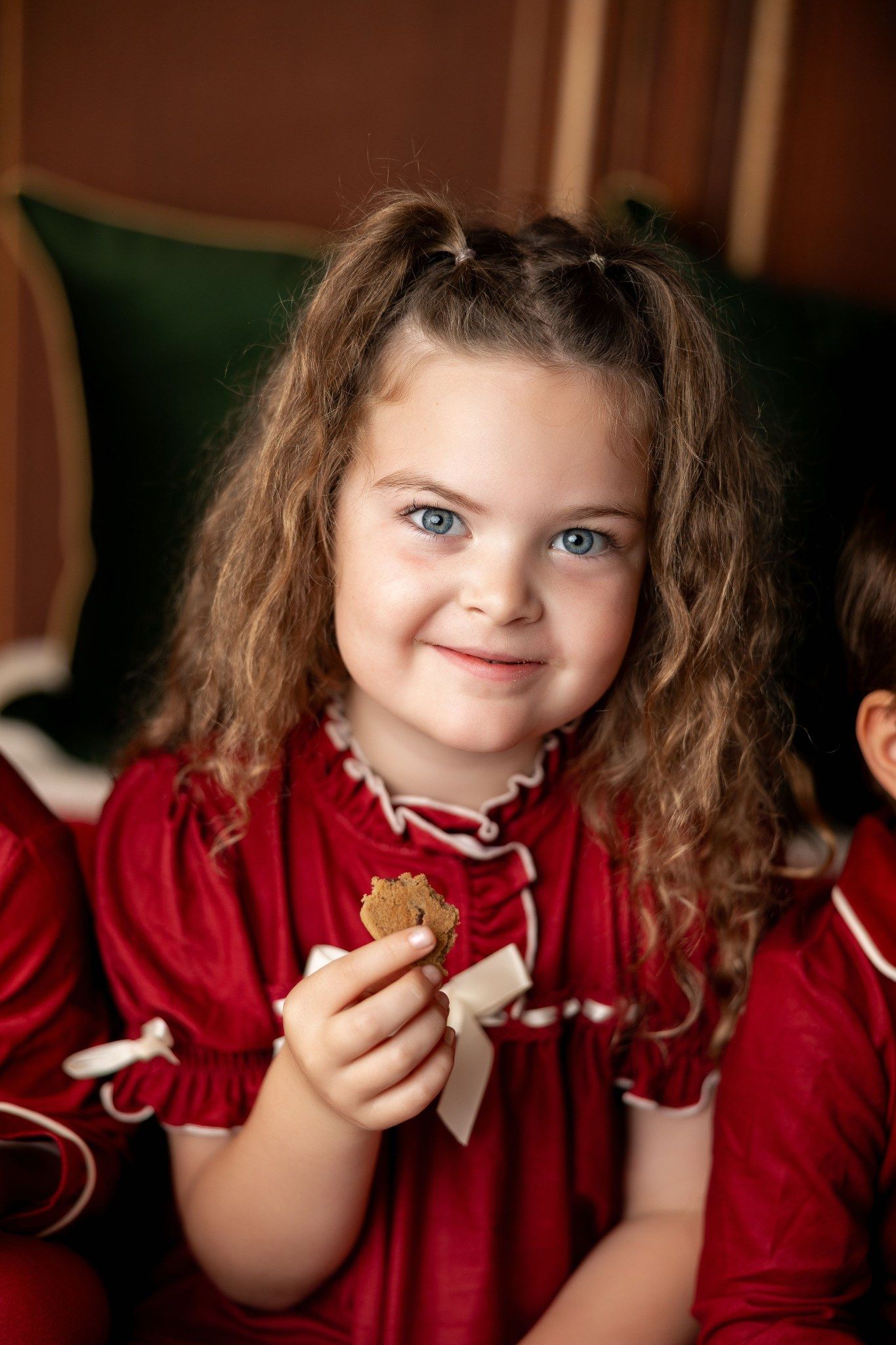 Smiling girl in a red velvet holiday dress holding a cookie at a 2025 Milk & Cookies Christmas mini session in Pacific Grove