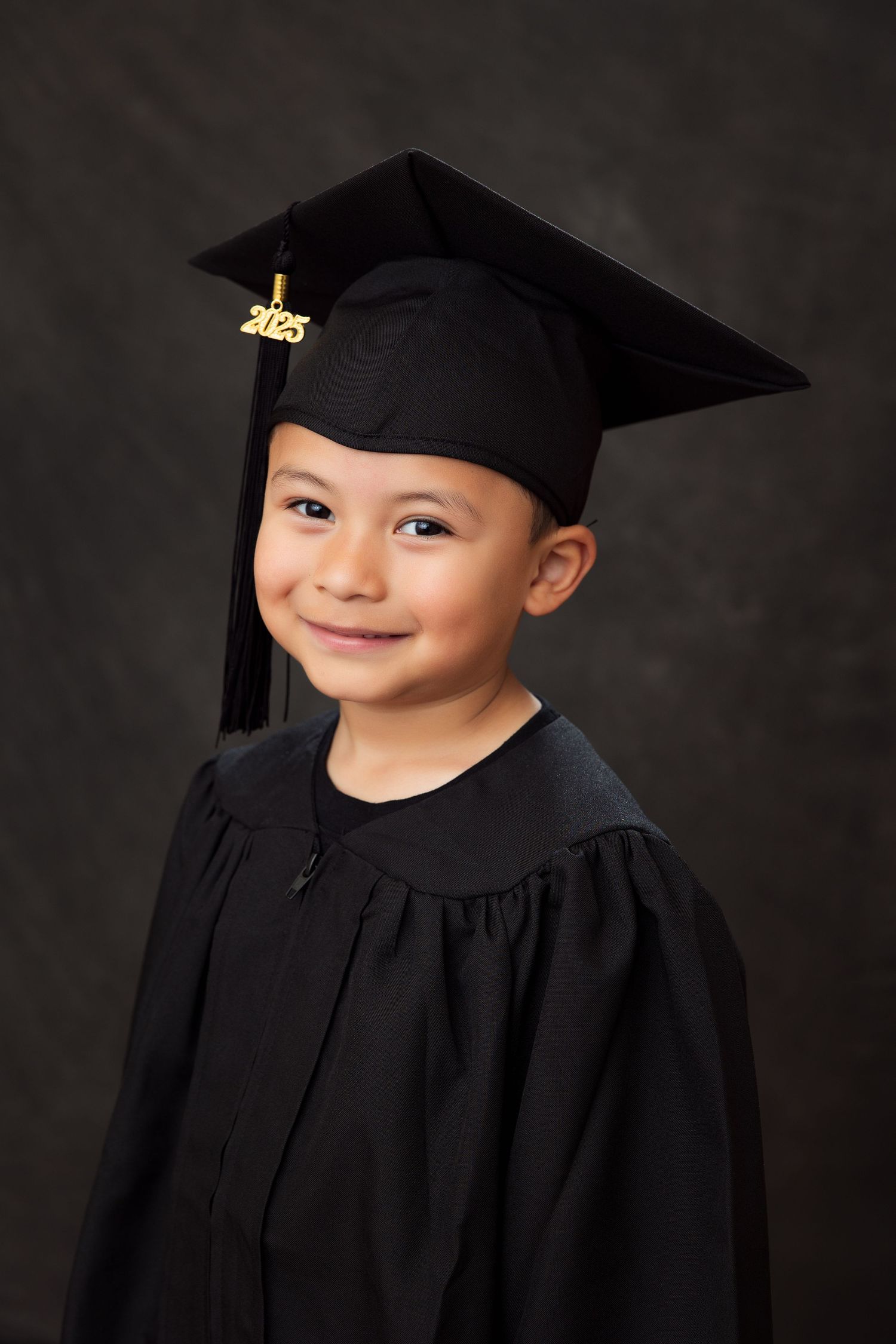 Studio portrait of a preschool graduate wearing traditional black cap and gown against dark background.