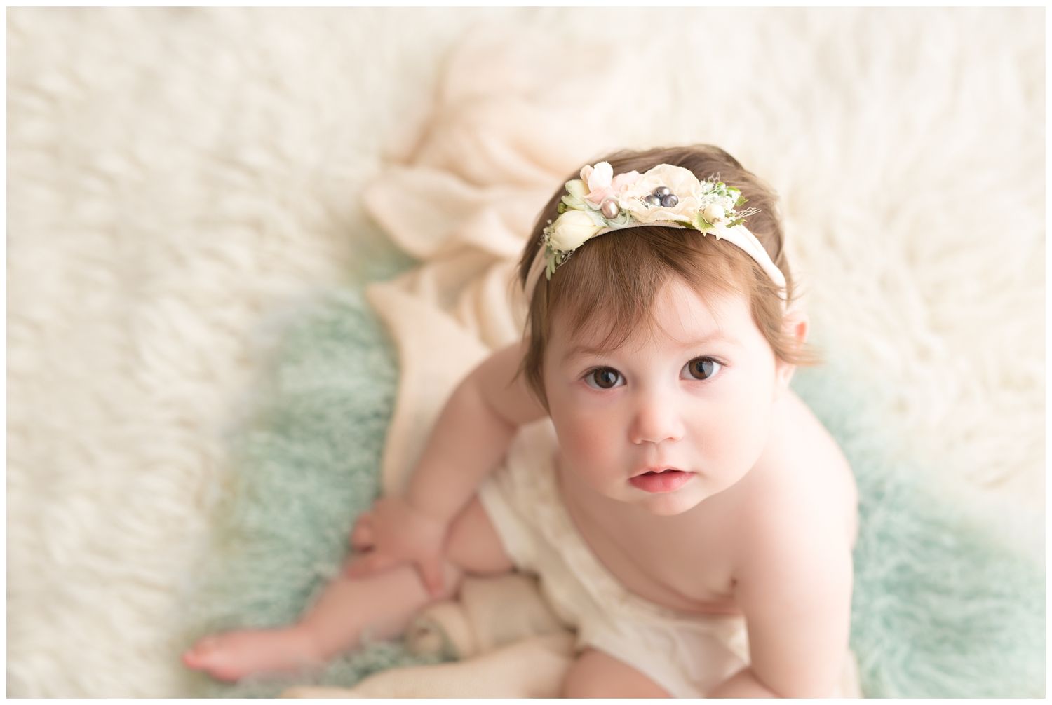 Baby wearing floral headband sits on mint green rug in soft pastel portrait setting.