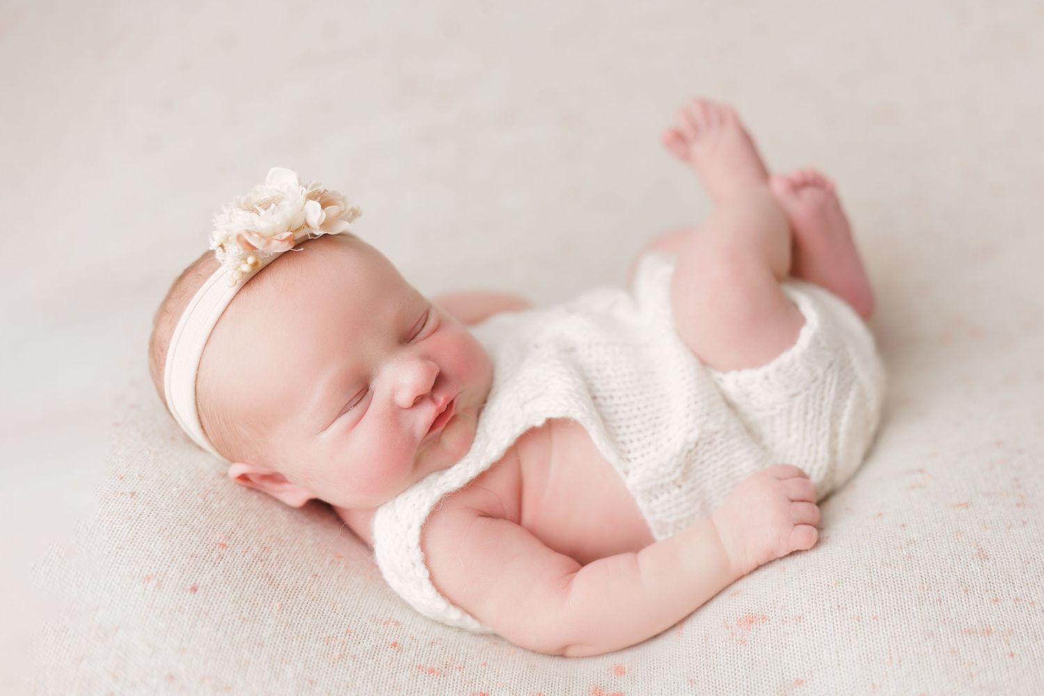 Sleeping newborn baby girl in white knit romper and floral headband on soft cream blanket.
