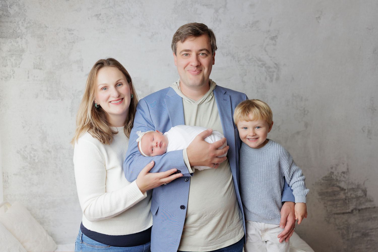 Happy family of four posing indoors, parents holding newborn baby with young blonde boy smiling together.