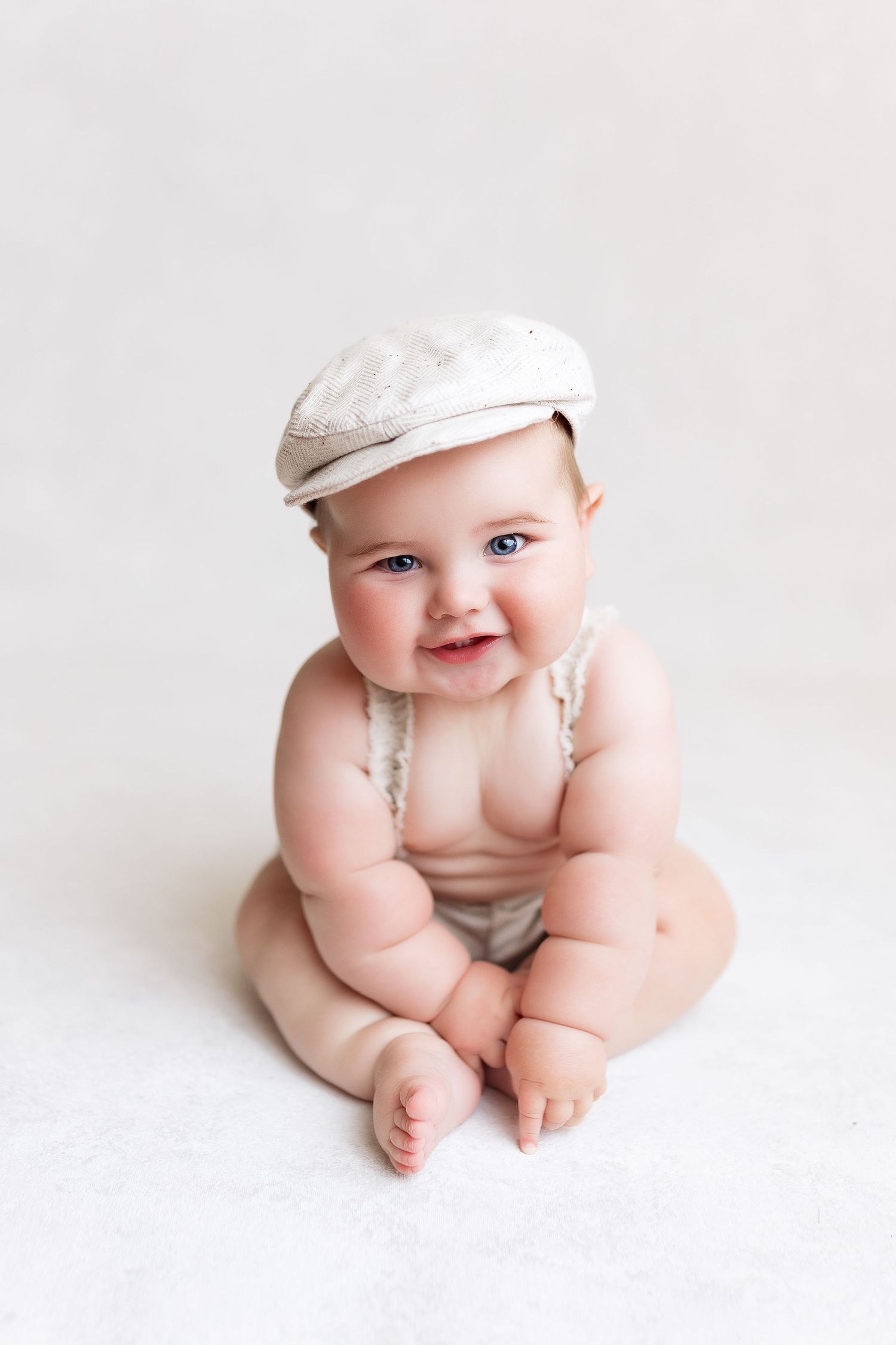 Smiling baby wearing a beige flat cap sits on white background in white romper showing chubby legs.