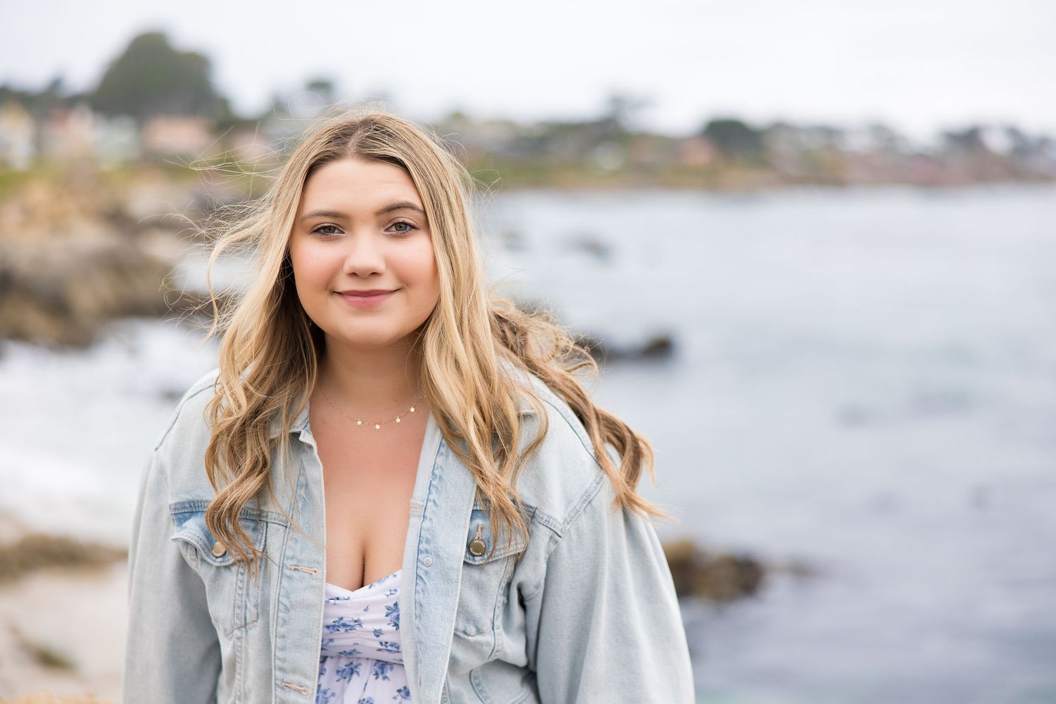 Young blonde woman in denim jacket smiling at rocky coastal beach with ocean and town in background.