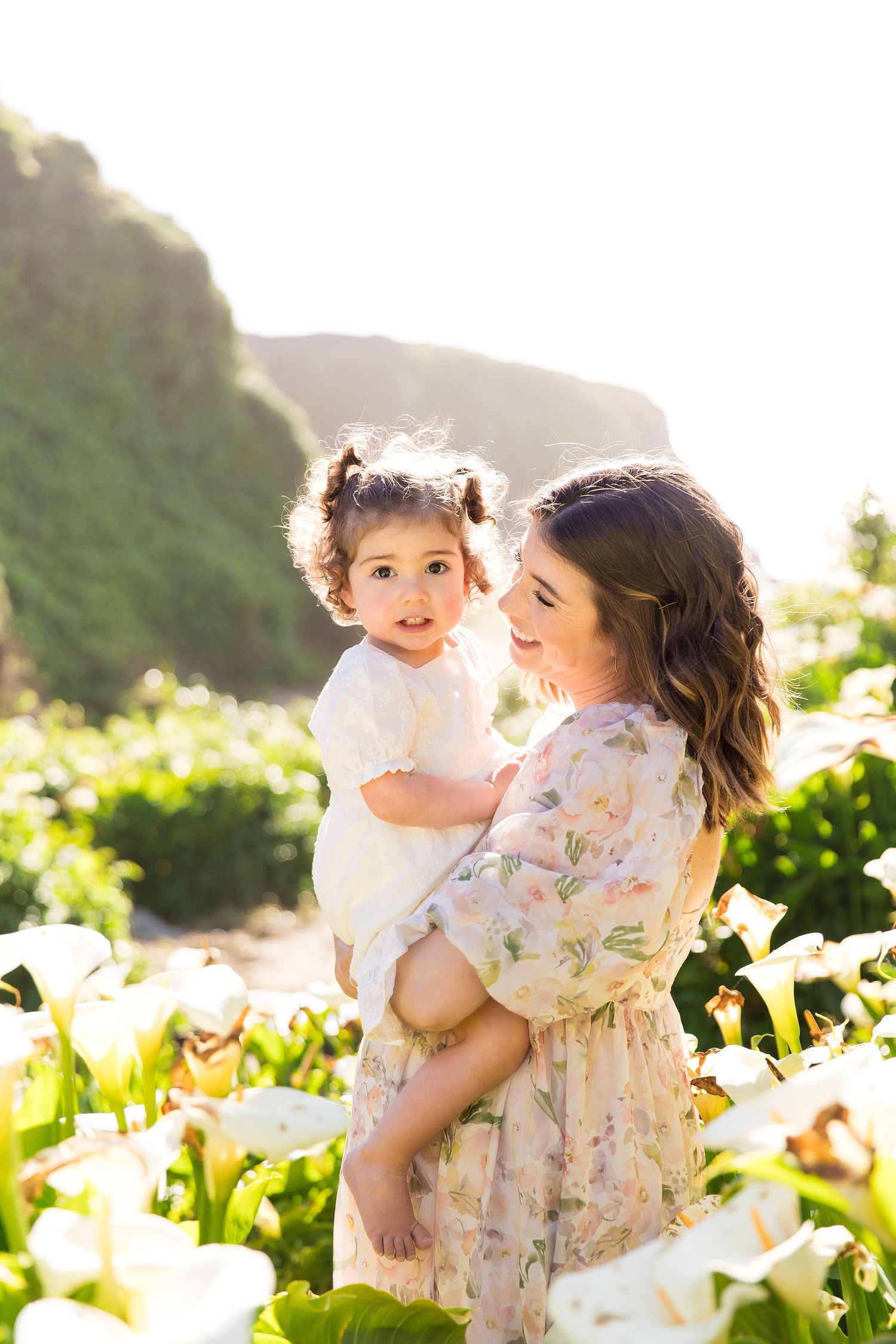 A mother in a floral dress holds her toddler daughter among white calla lily valley Big Sur.