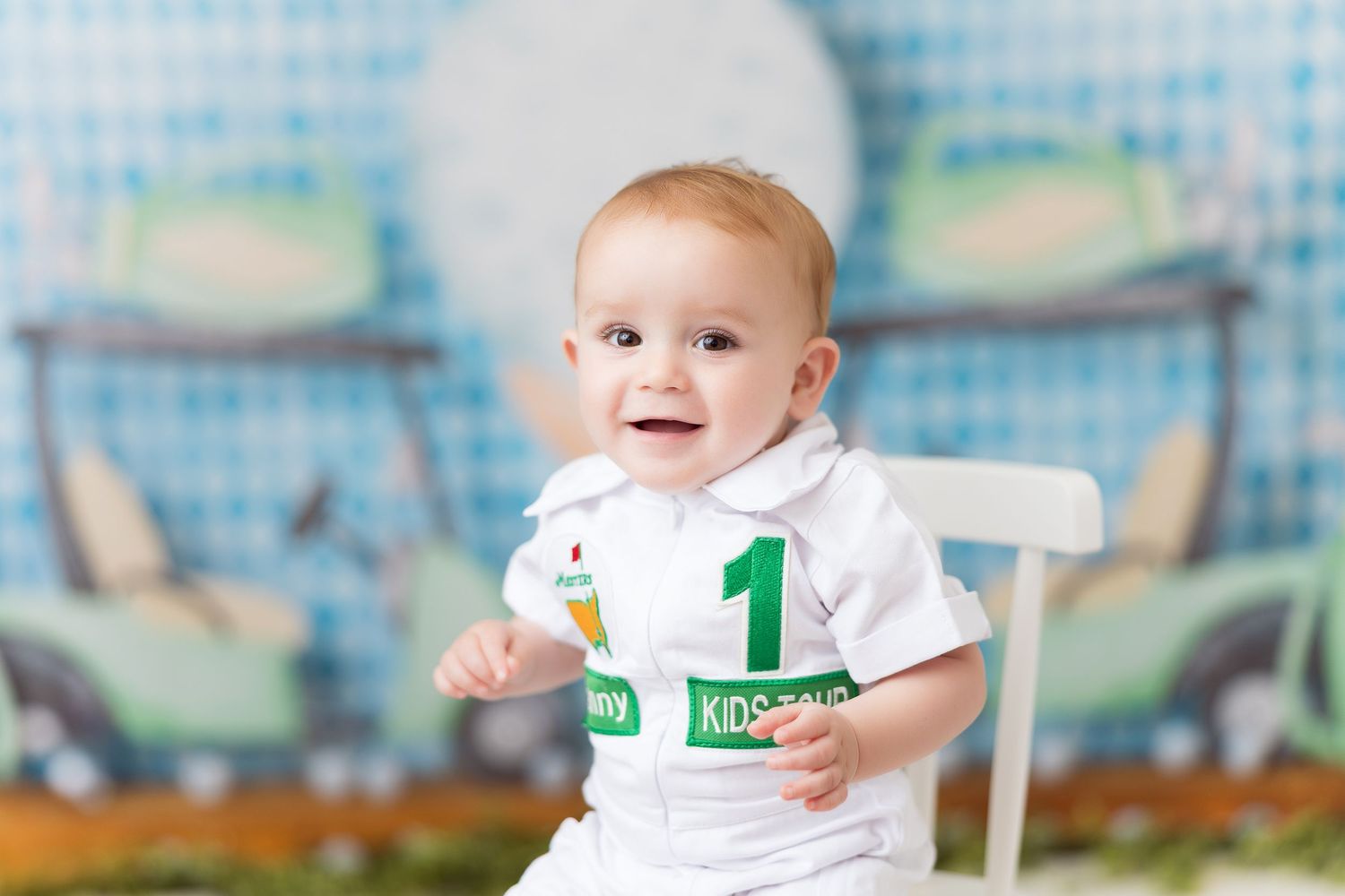 Baby wearing number one shirt sits against blue and white checkered backdrop with birthday setup.