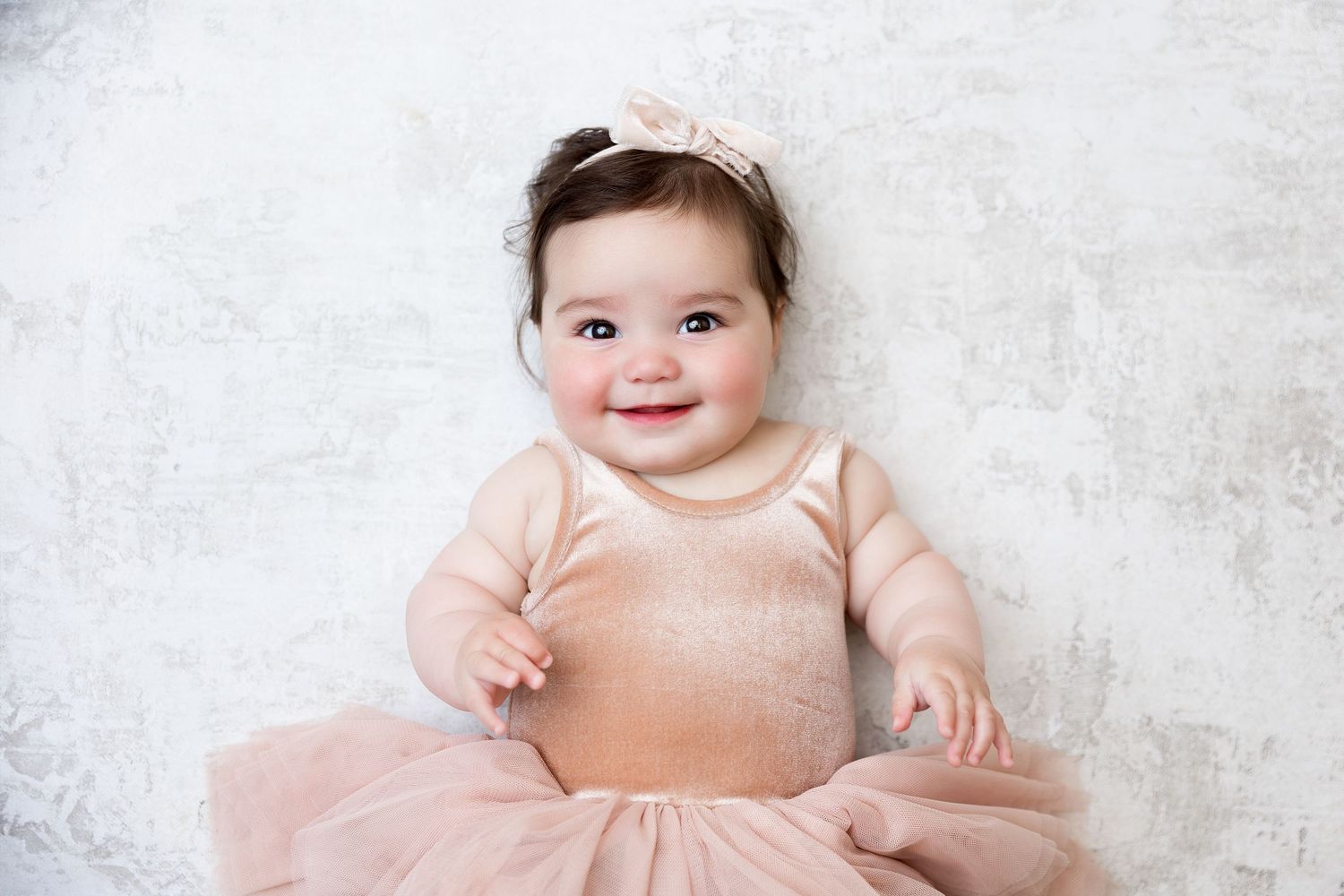 Baby in peach tulle dress lies on white textured background wearing small hair bow.
