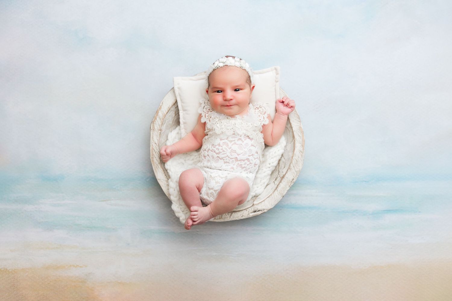 Baby wearing white outfit lying in rustic round basket against dreamy beach backdrop.