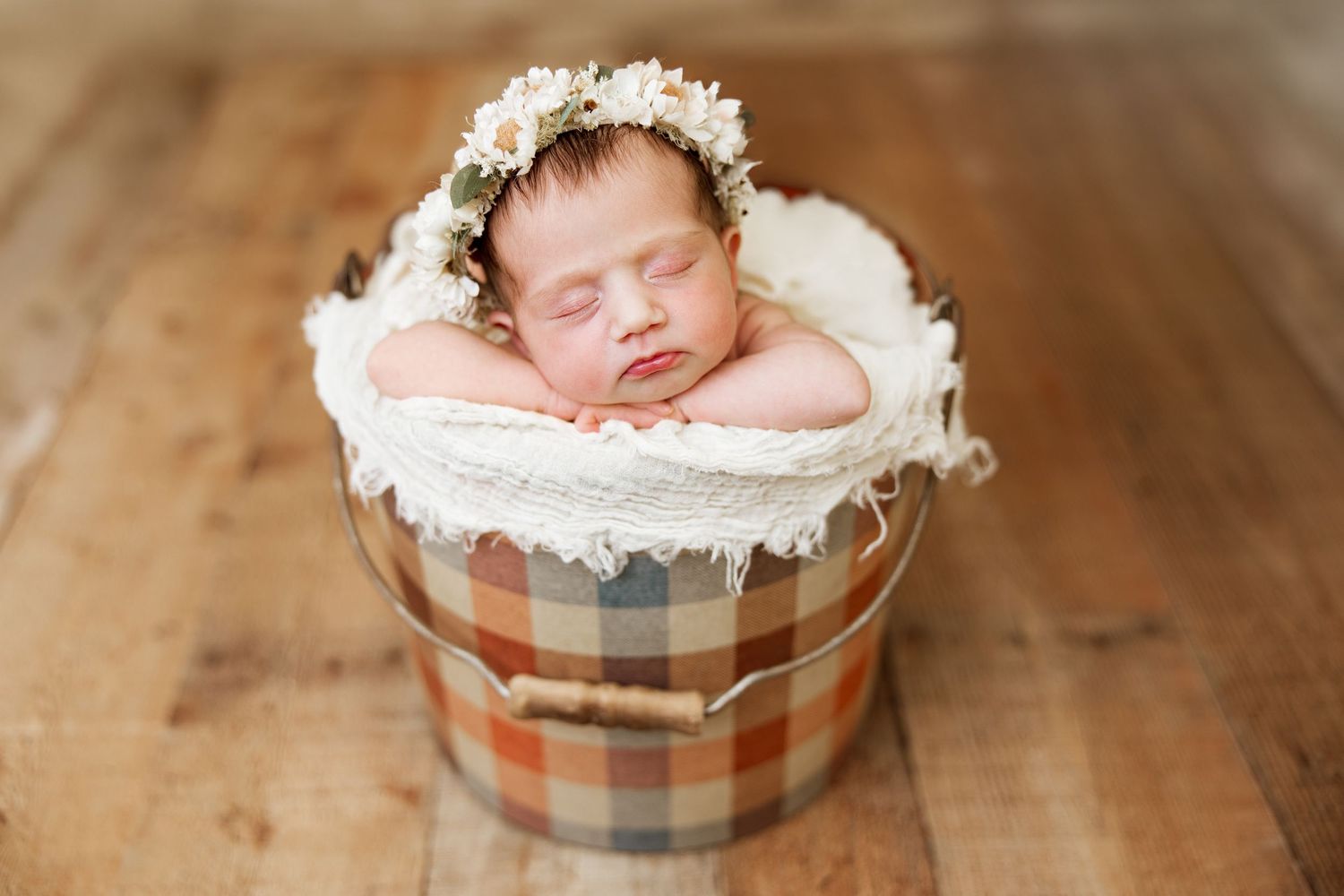Sleeping newborn posed in round wooden container with soft white padding and floral crown.