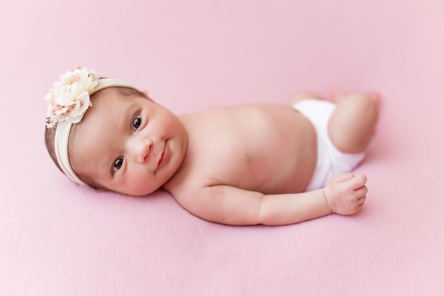 Baby wearing floral headband lying on pink background.