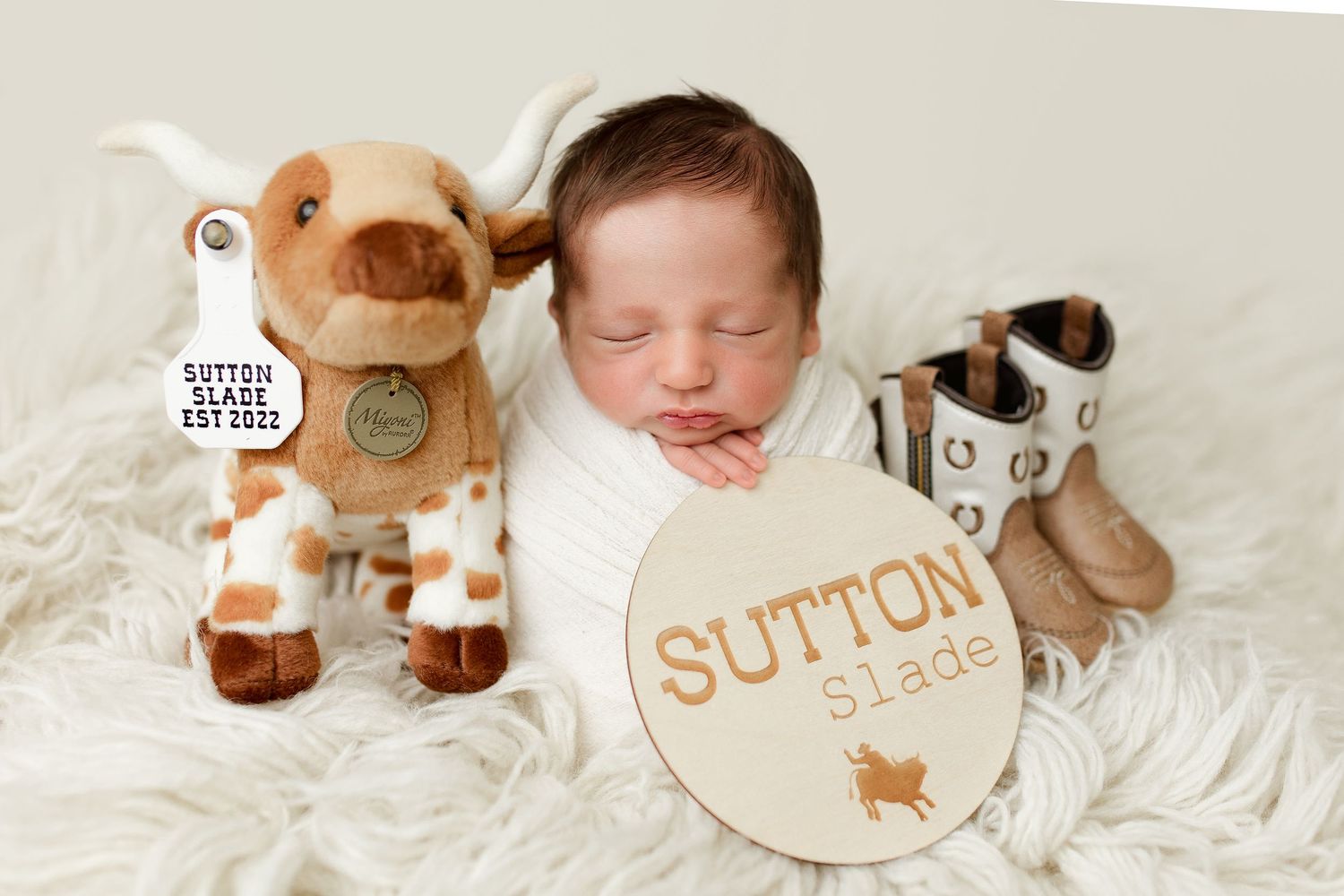 Newborn baby sleeping peacefully on white fur with plush deer toy and wooden name sign.