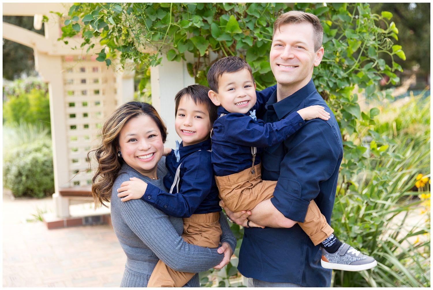 A happy family of four poses together outdoors in a garden setting on a sunny day.