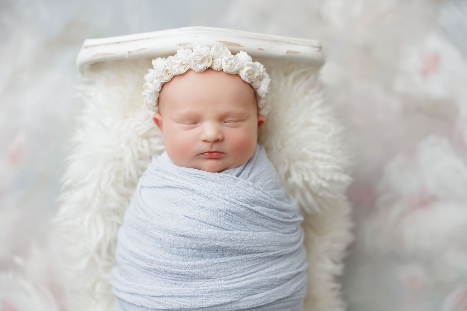 Newborn baby swaddled in light blue wrap, wearing floral headband, sleeping peacefully in white wooden bed with fluffy fur.