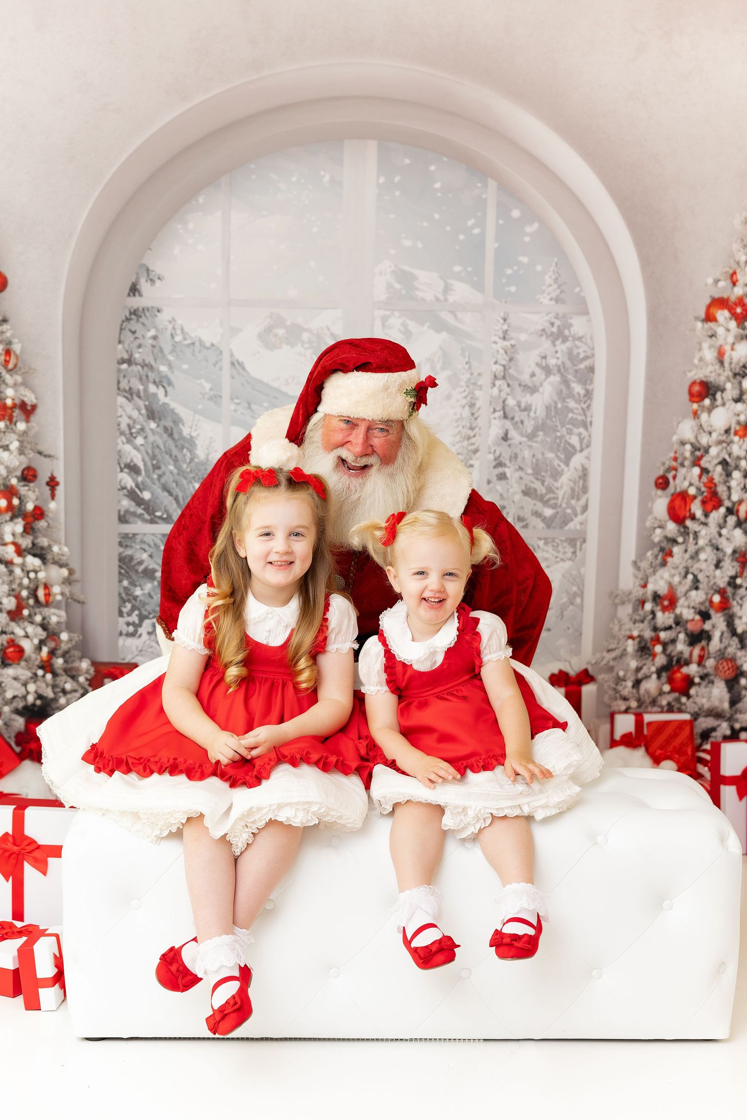 A festive Christmas photo shoot with two children and Santa against a snowy white backdrop with decorated trees.