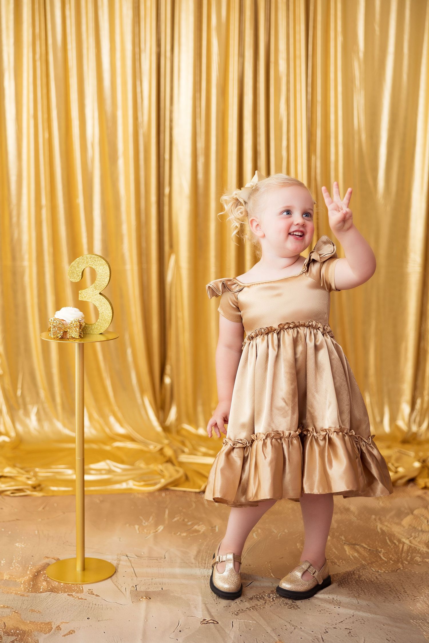 Young child in gold party dress poses happily with peace sign near gold number 3 decoration against golden curtain backdrop.