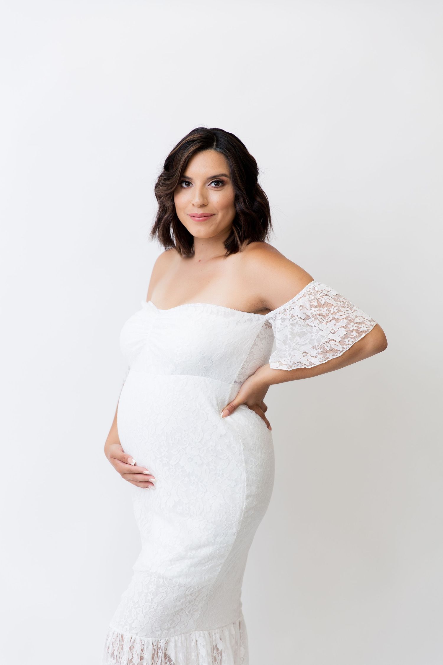 Elegant maternity portrait in white lace off-shoulder dress against white background.