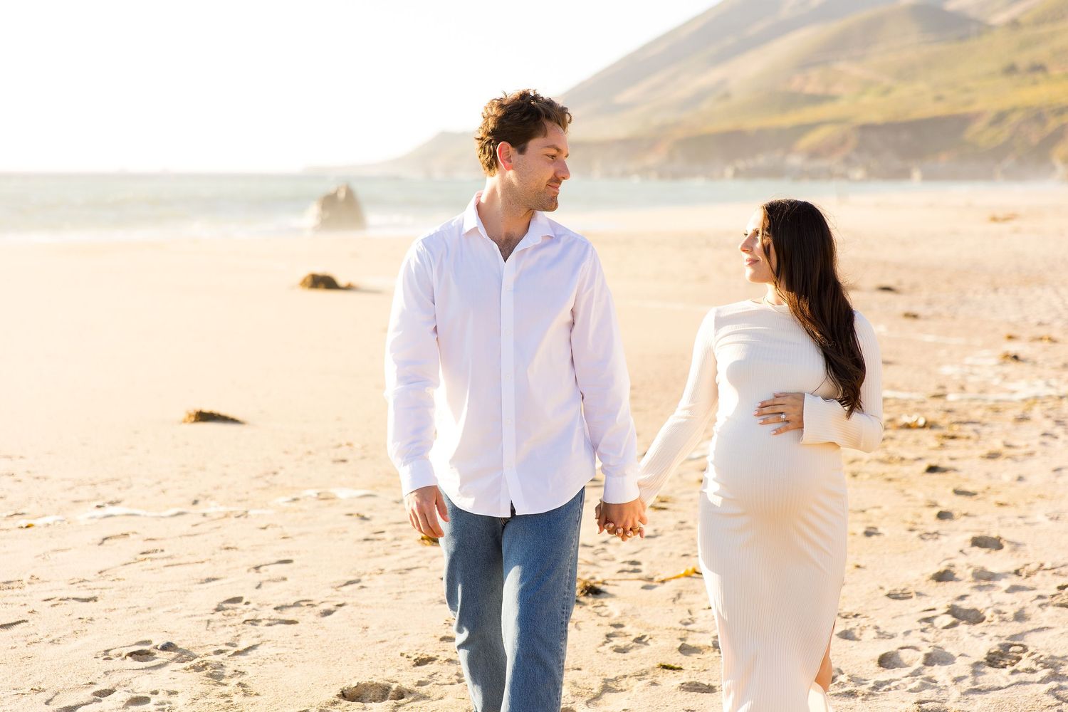 Couple walking hand in hand on a sandy beach with mountains in the background.