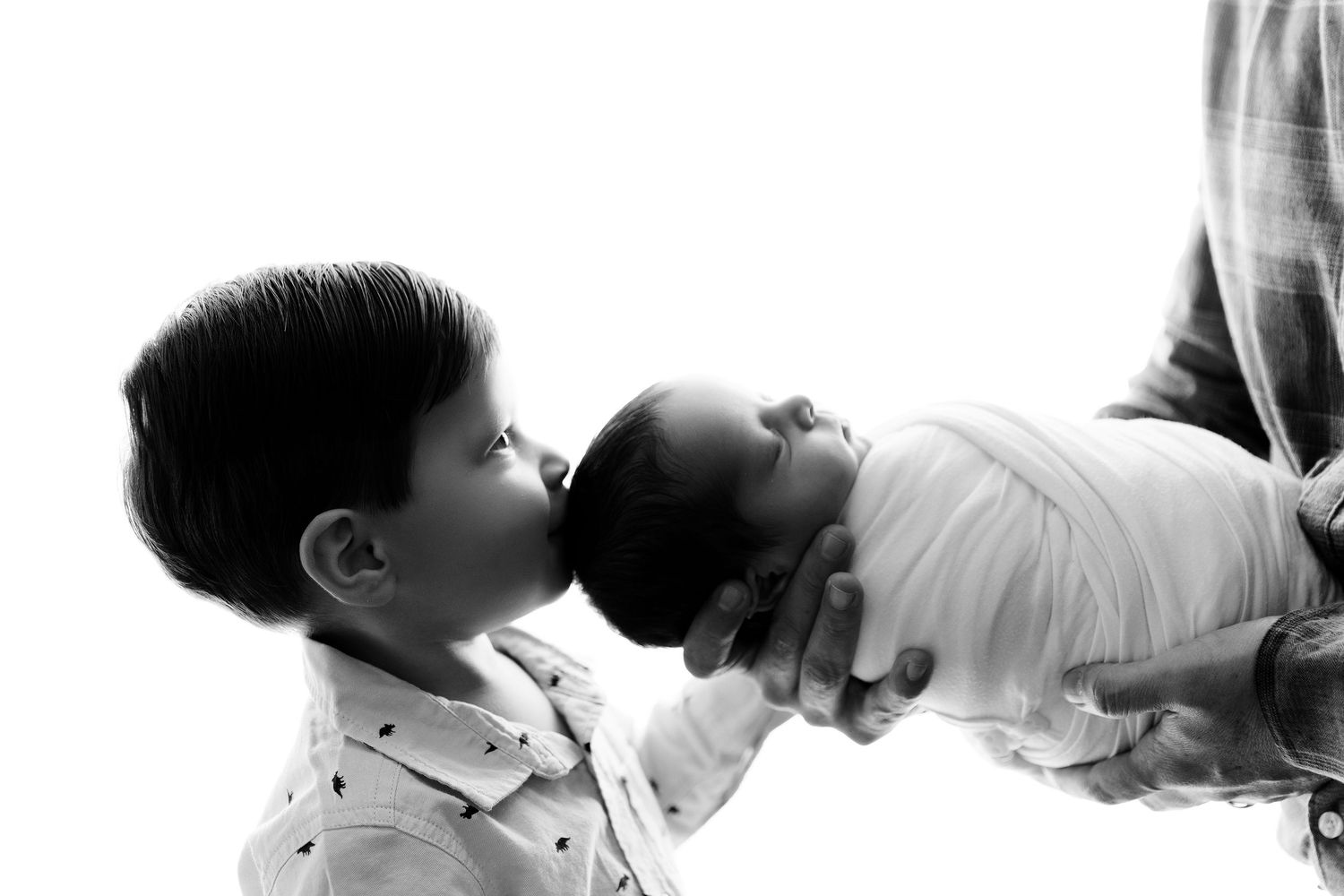 Black and white portrait of sibling kissing newborn baby wrapped in white fabric.