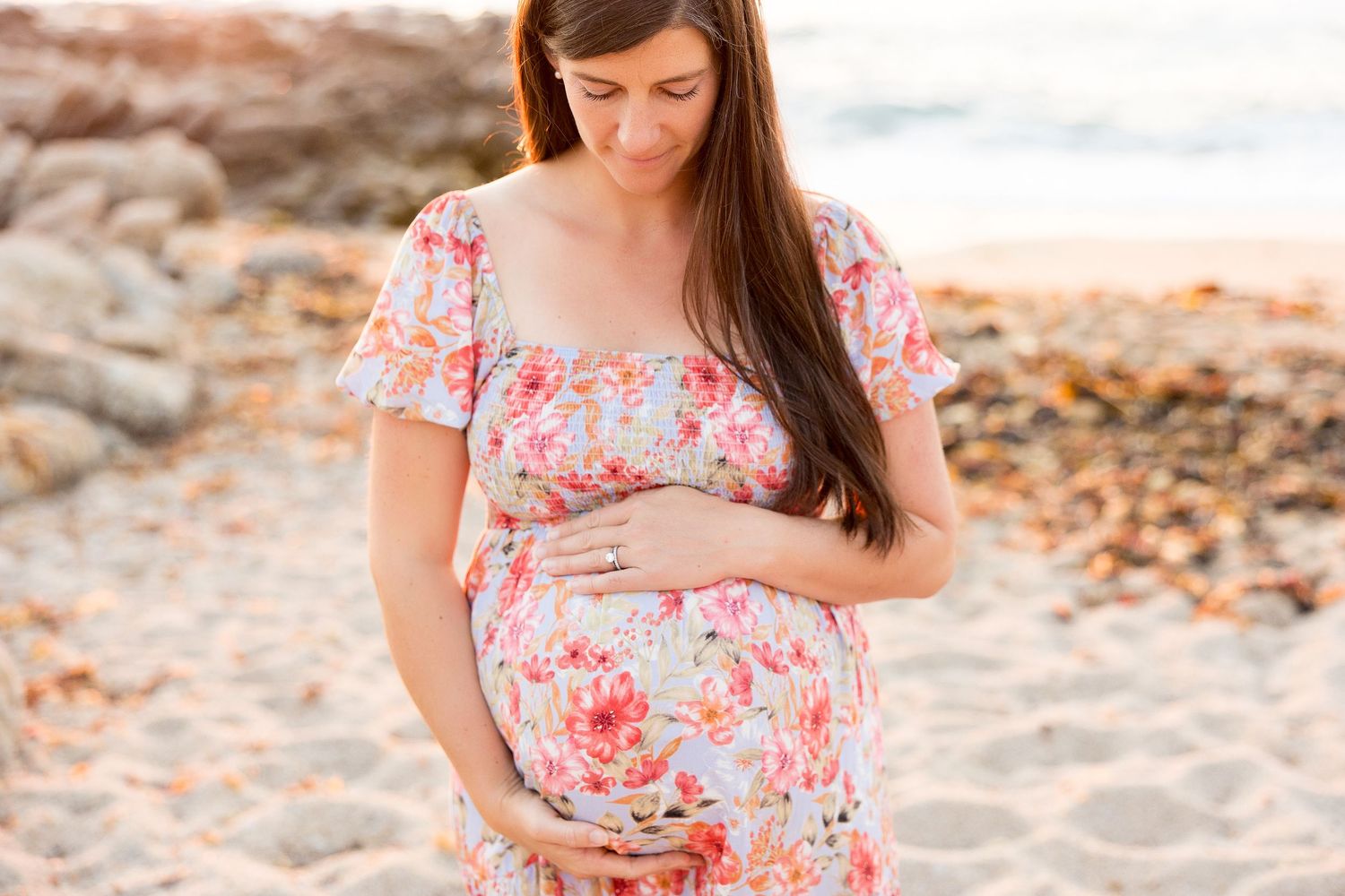 A woman in a floral dress cradles her baby bump on a sandy beach at sunset.