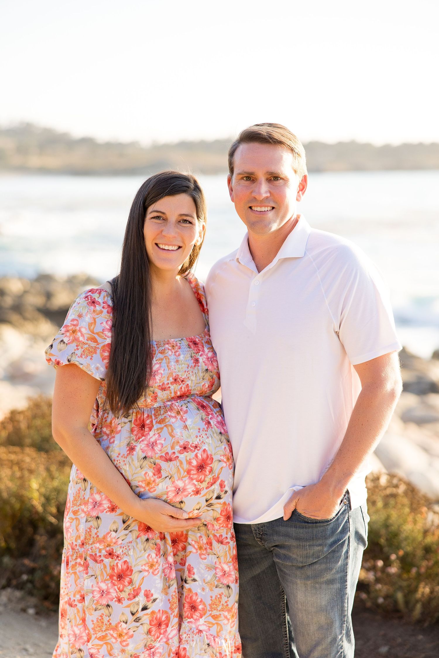 A couple poses together on a rocky beach during golden hour wearing white and floral outfits.