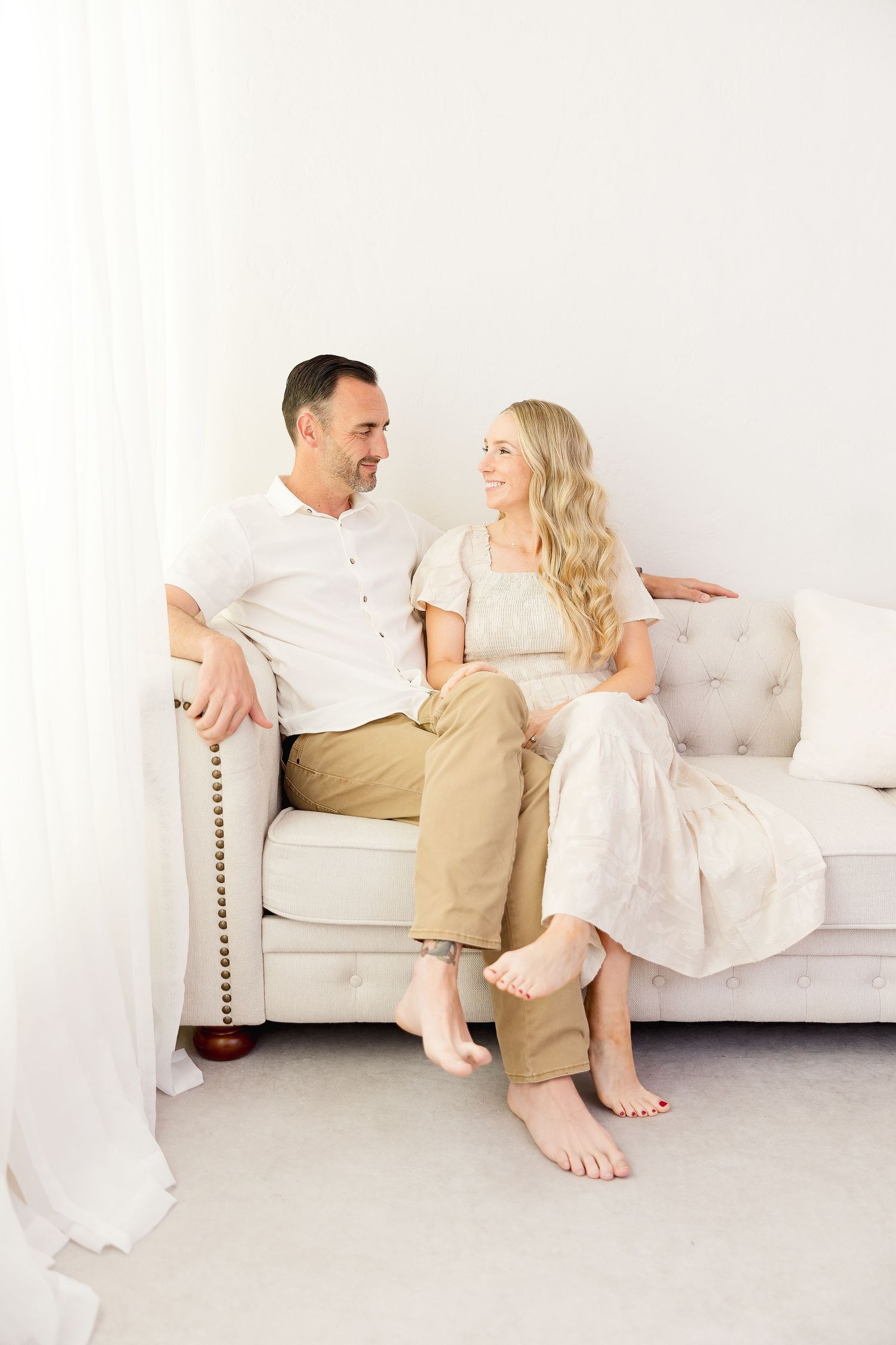 Couple in white and beige outfits relaxing and laughing together on a light colored sofa in a bright minimalist setting.