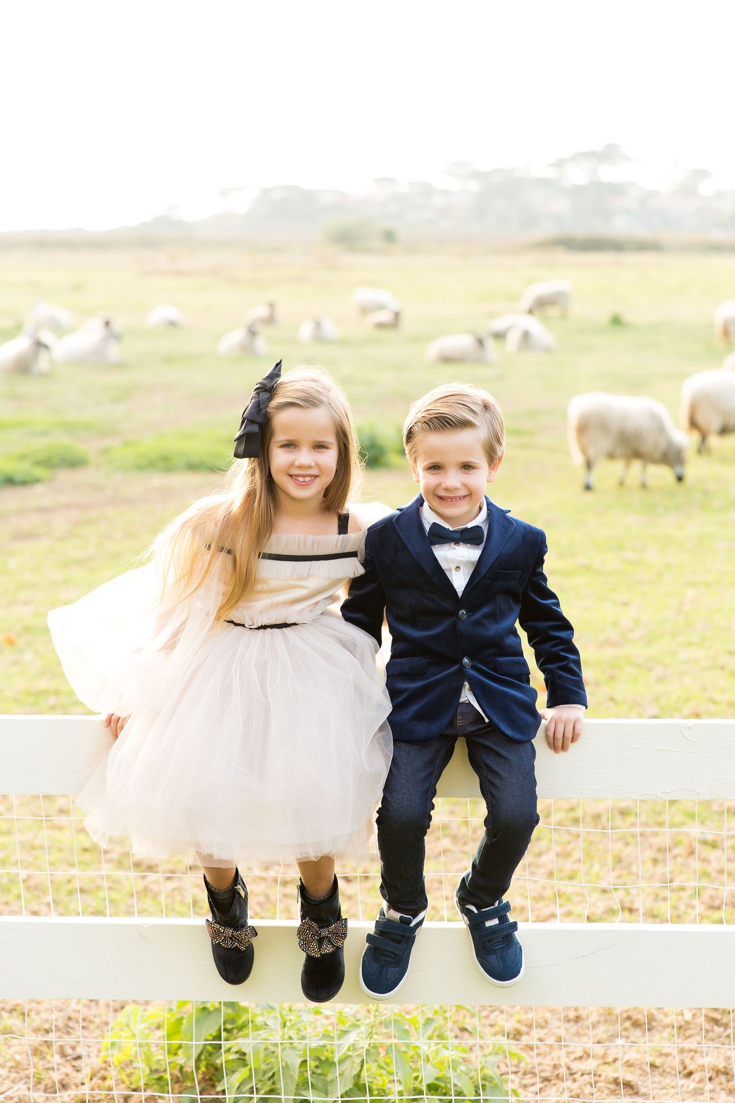 Children in formal attire sitting on white fence with sheep grazing in pasture behind them.