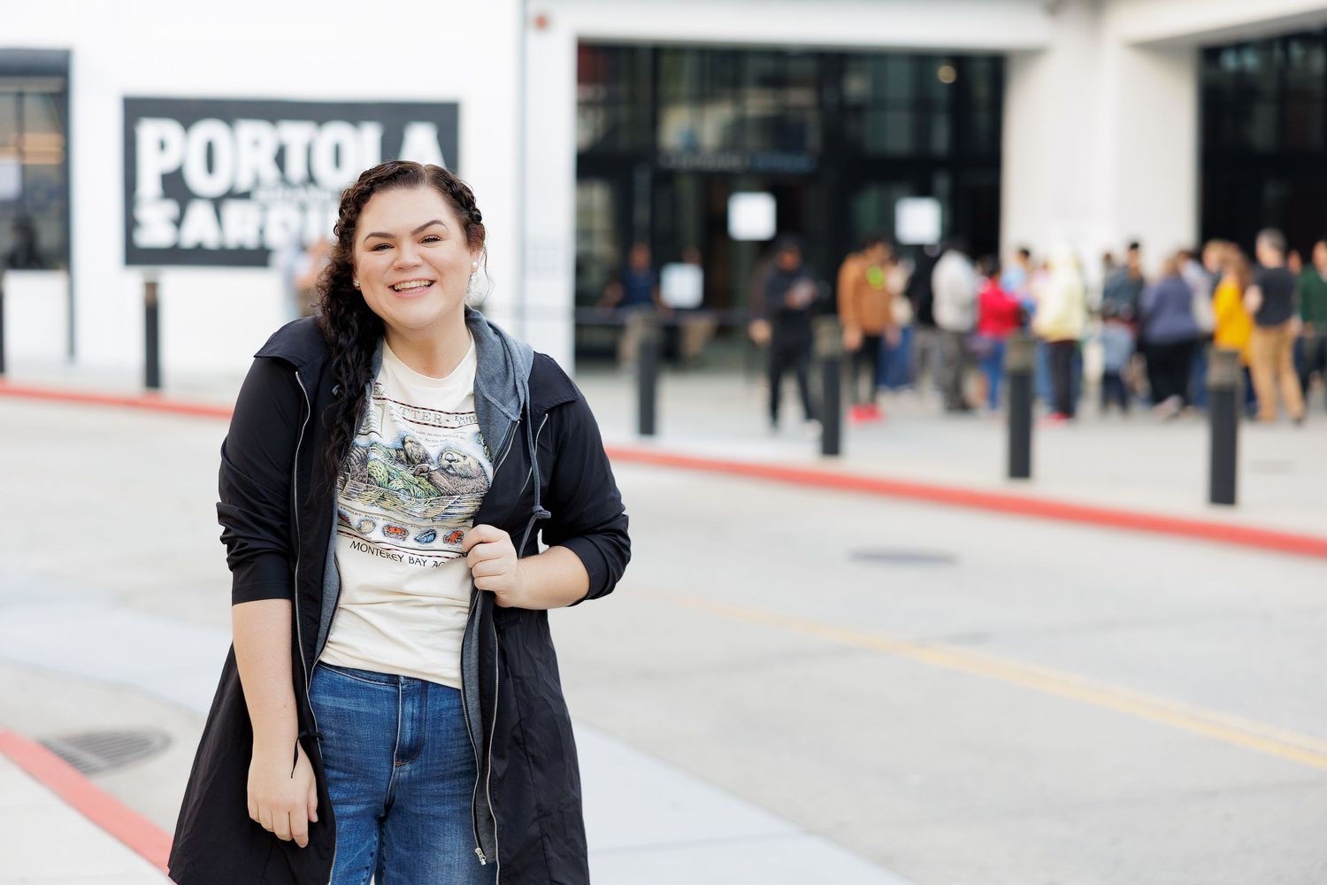 A person in a black jacket and blue jeans stands smiling in front of an outdoor storefront.