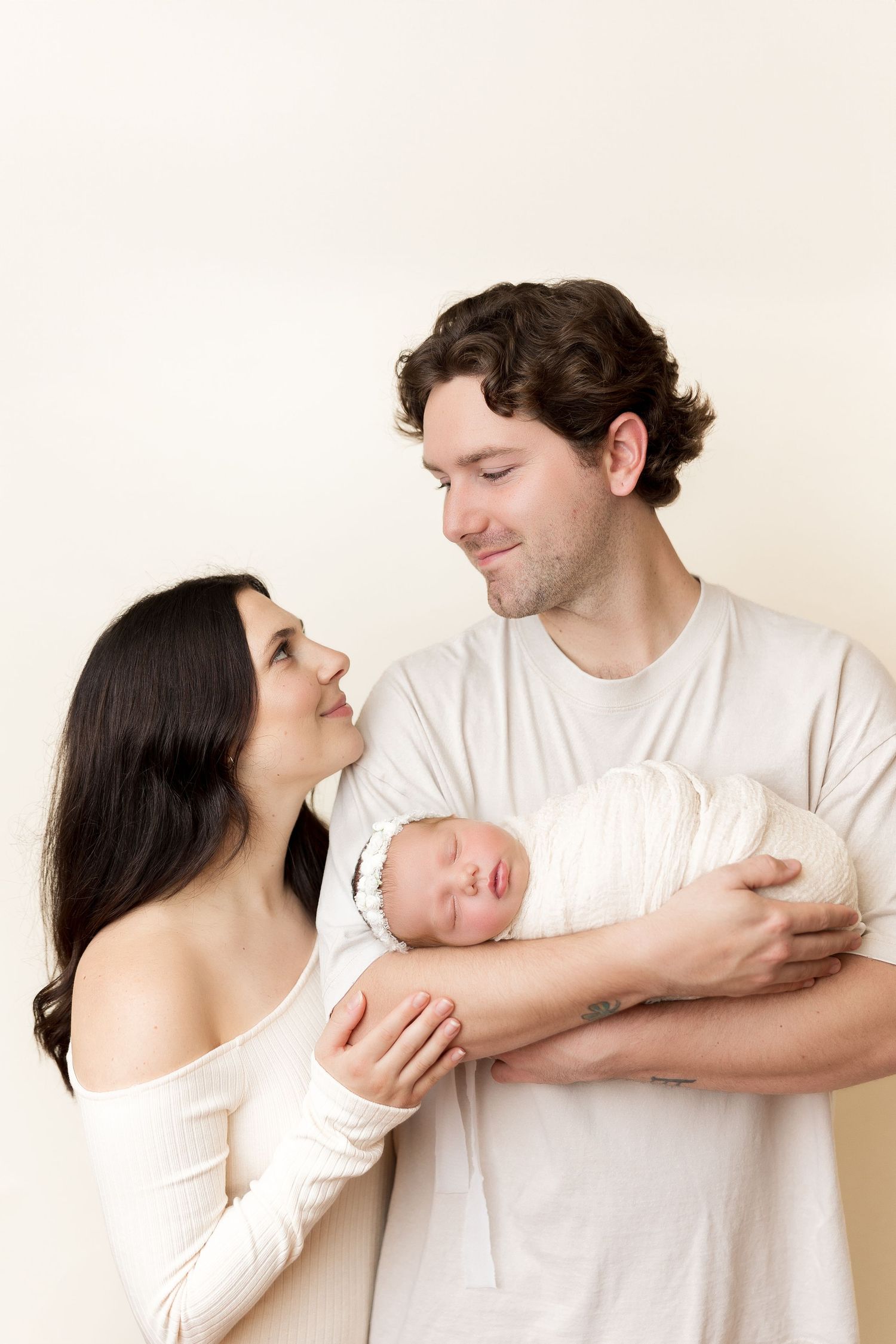 Parents share tender moment while holding their sleeping newborn wrapped in white.