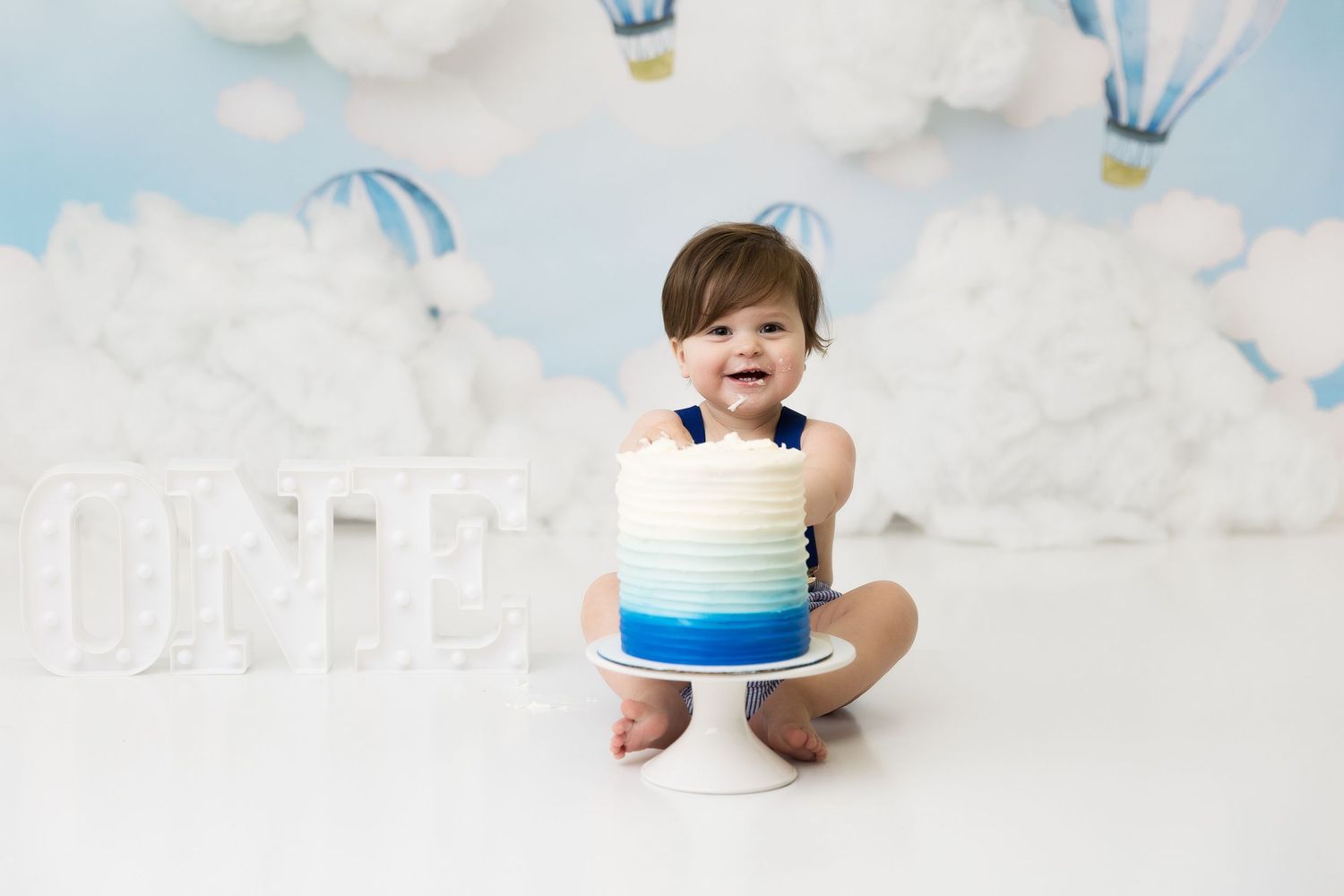 Happy baby posing with blue and white cake against hot air balloon backdrop.