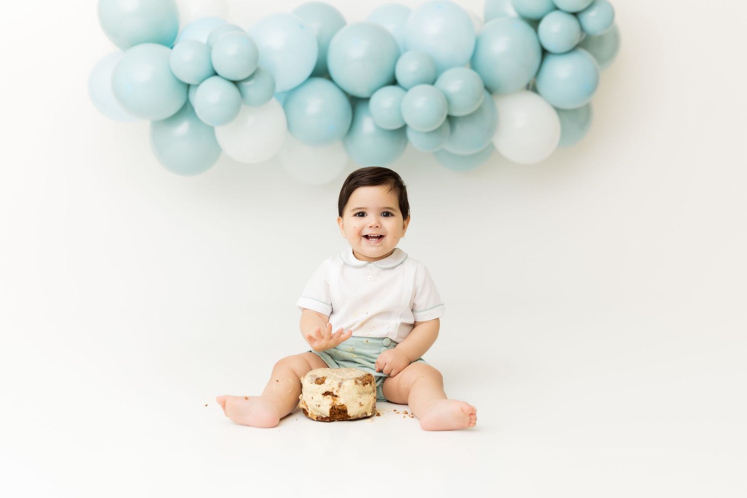 Happy baby sitting with cake against pastel blue balloon garland.