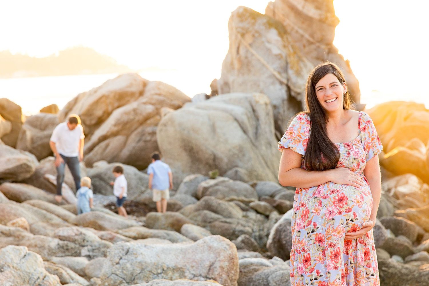 Maternity photo session on rocky beach with floral dress and warm sunset lighting.