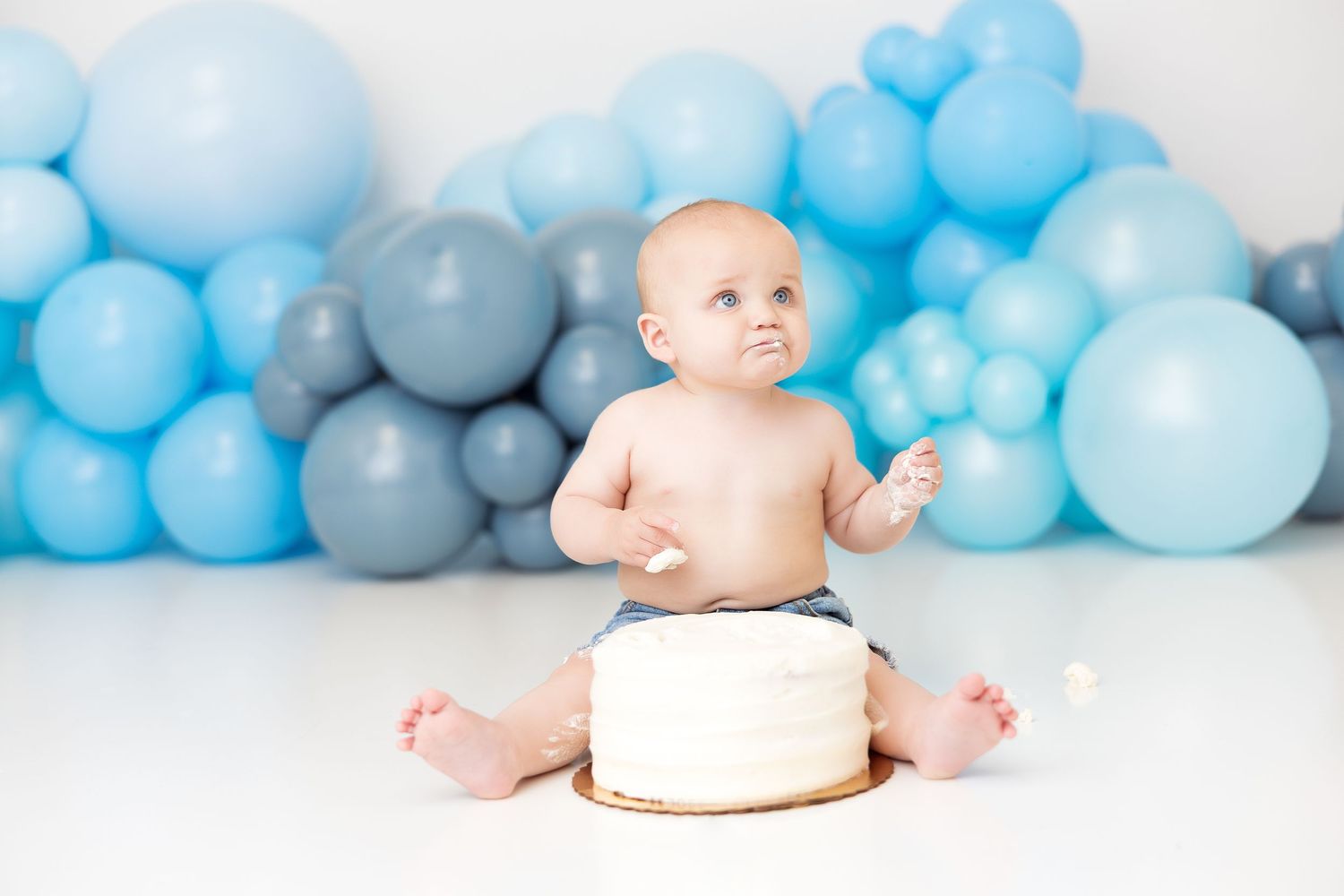 A small baby sits in front of blue and gray balloon decorations with a white cake for a cake smash photo session.
