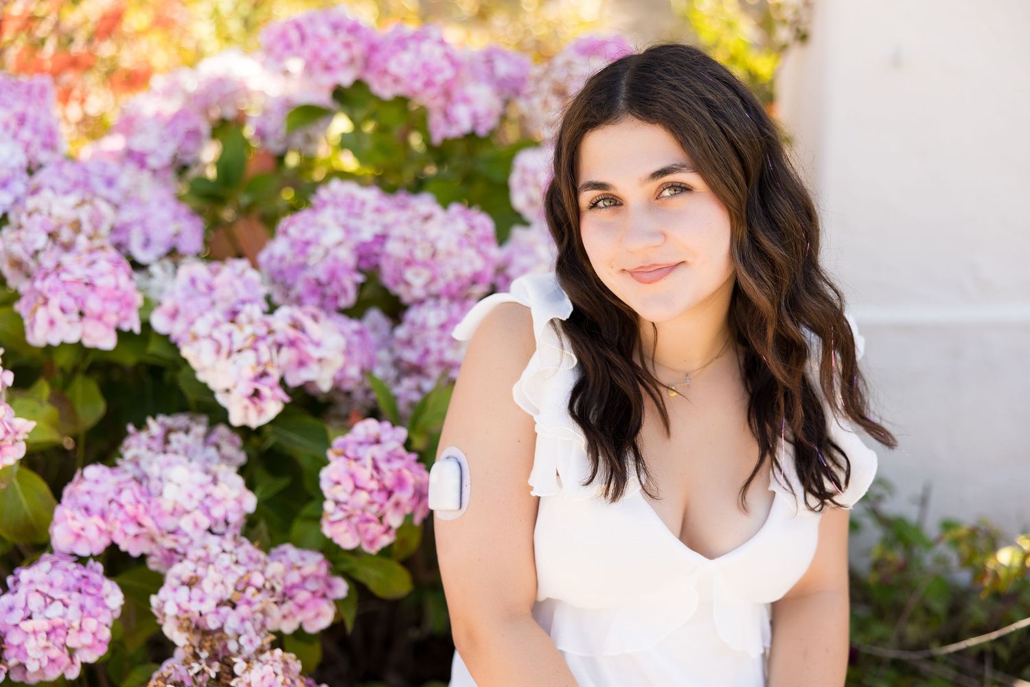 Young woman in white dress sitting near blooming pink hydrangeas in a sunny outdoor garden setting.